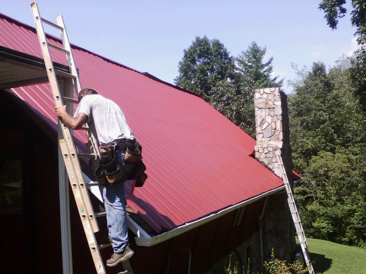 Roofer on a ladder beside a red metal roof and stone chimney — Soffit & Fascia Repair by Roofing Brothers in Big Stone Gap